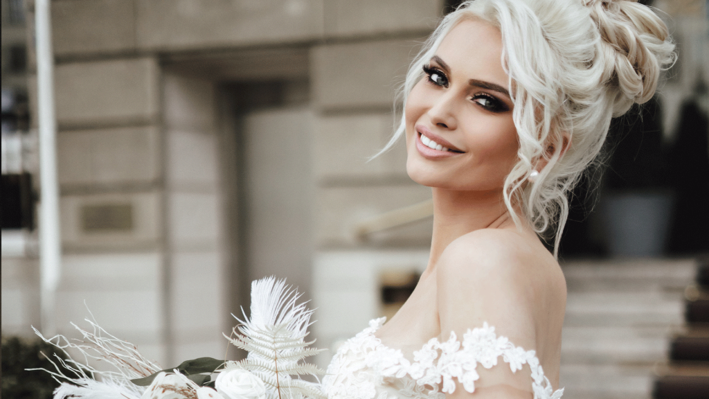 Fröhliche Braut mit lockerer Hochsteckfrisur und glamurösem Makeup in seitlicher Ansicht mit Blumenstrauss