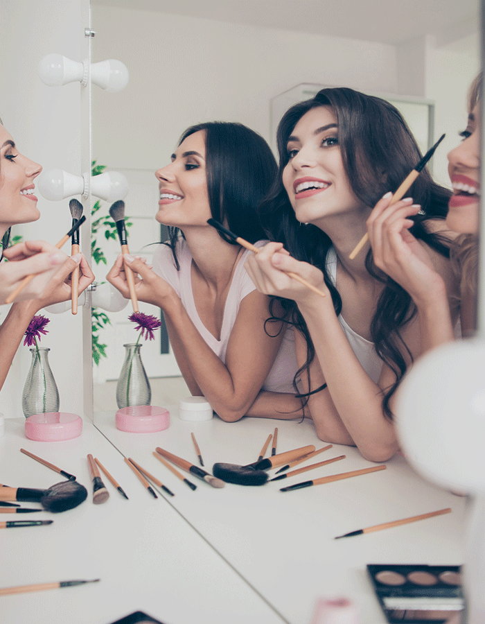 Gruppe junger Frauen sitzt lachend vor dem Spiegel und schminkt sich gemeinsam mit Pinseln